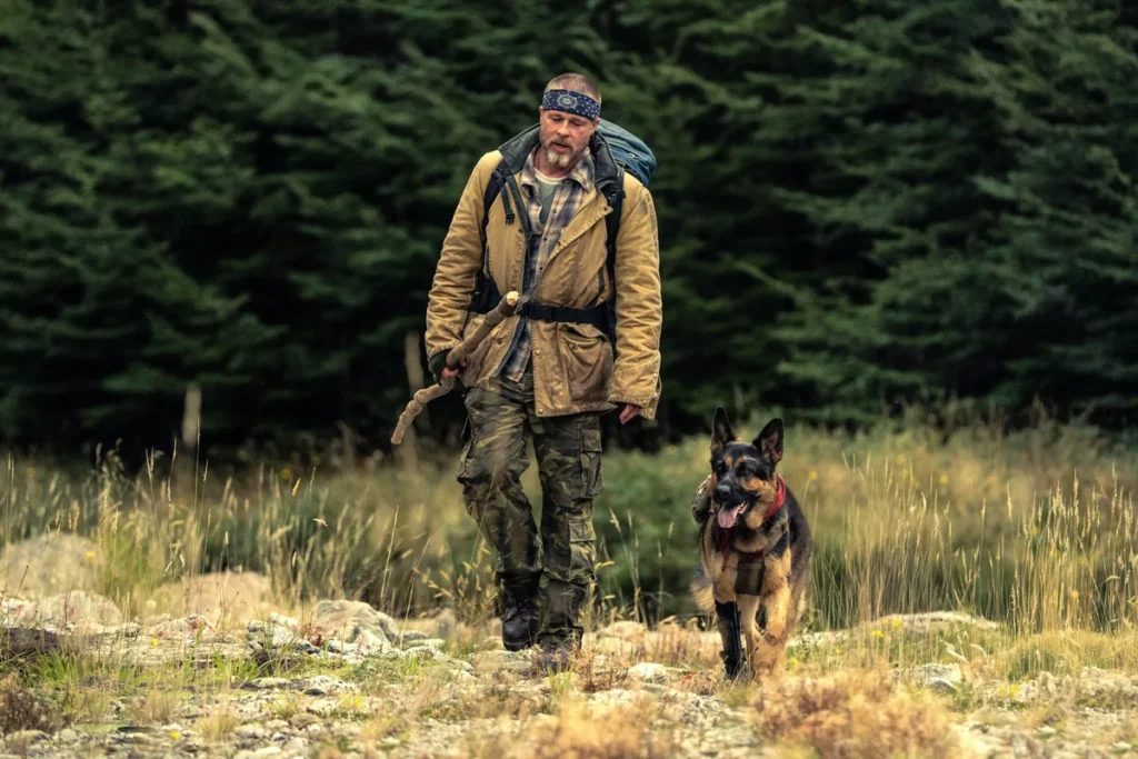 Heart of the Beast (2026): Brad Pitt as James Belmont walks through a grassy clearing beside Odin, a German Shepherd with a prosthetic front leg and a red bandana